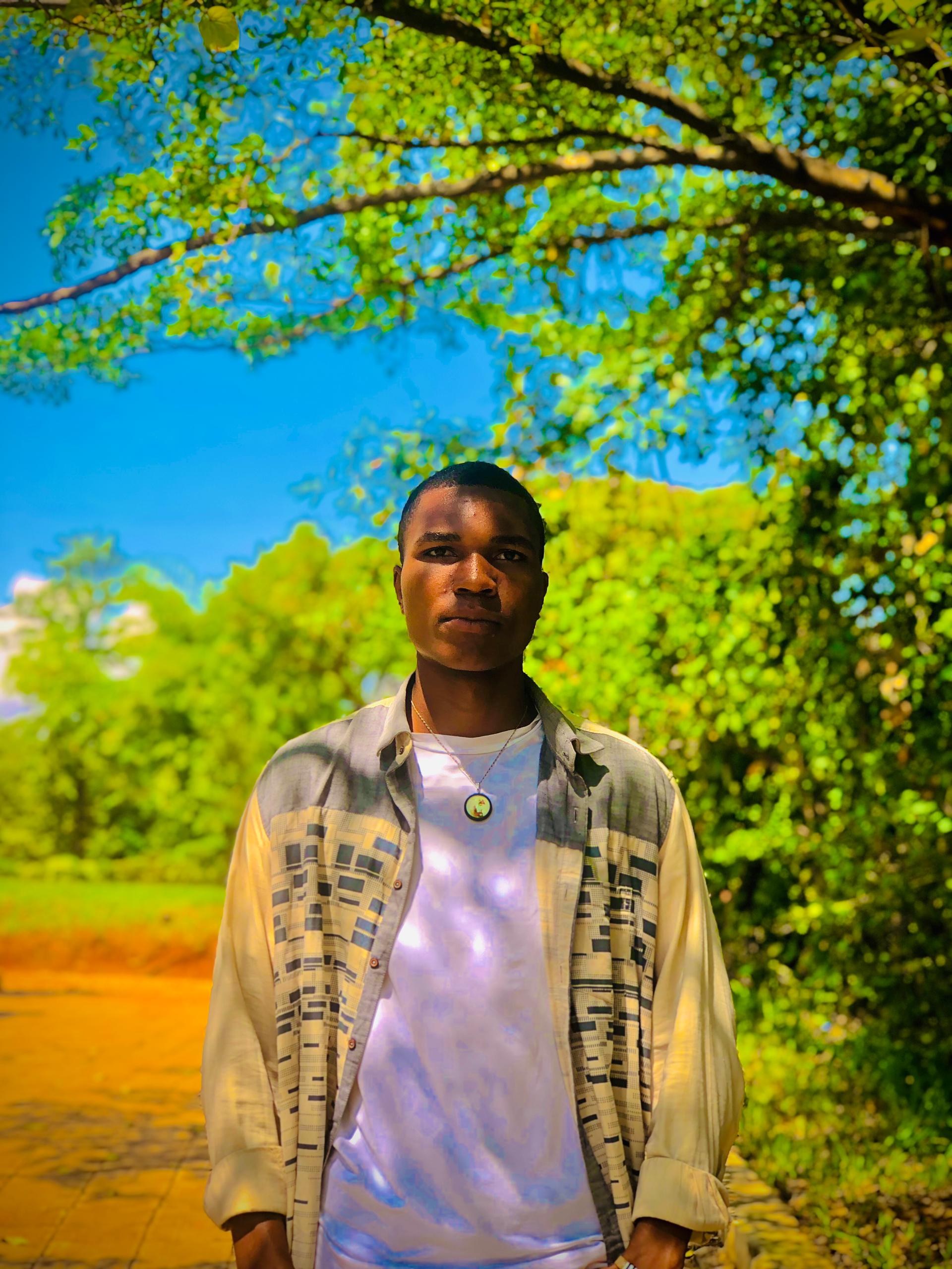 Person wearing a patterned overshirt and white T-shirt beneath leafy trees against a blue sky