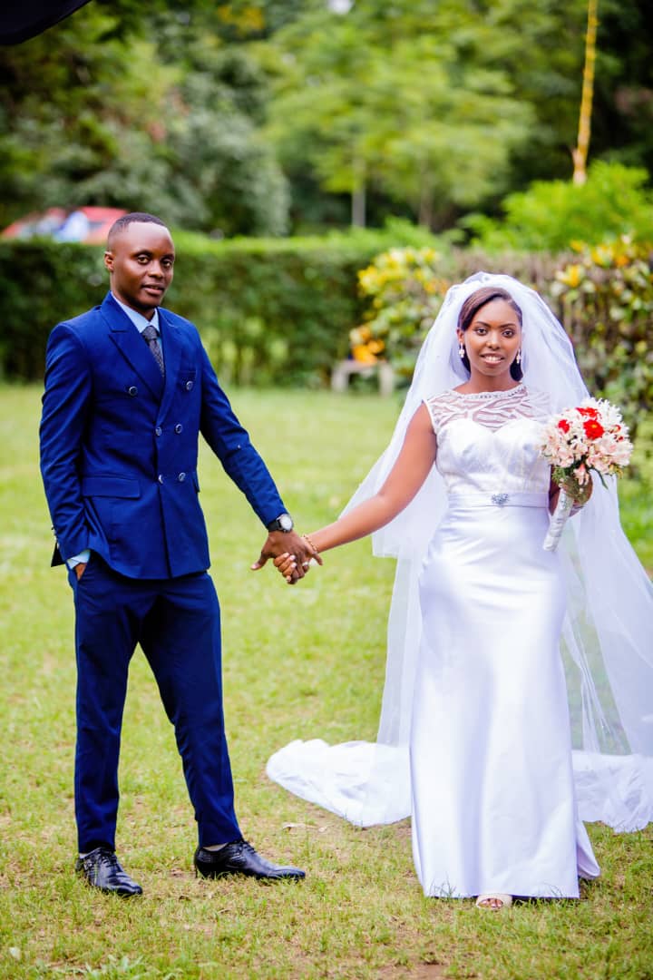 Bride in a white gown holding hands with a groom in a blue suit on a garden lawn