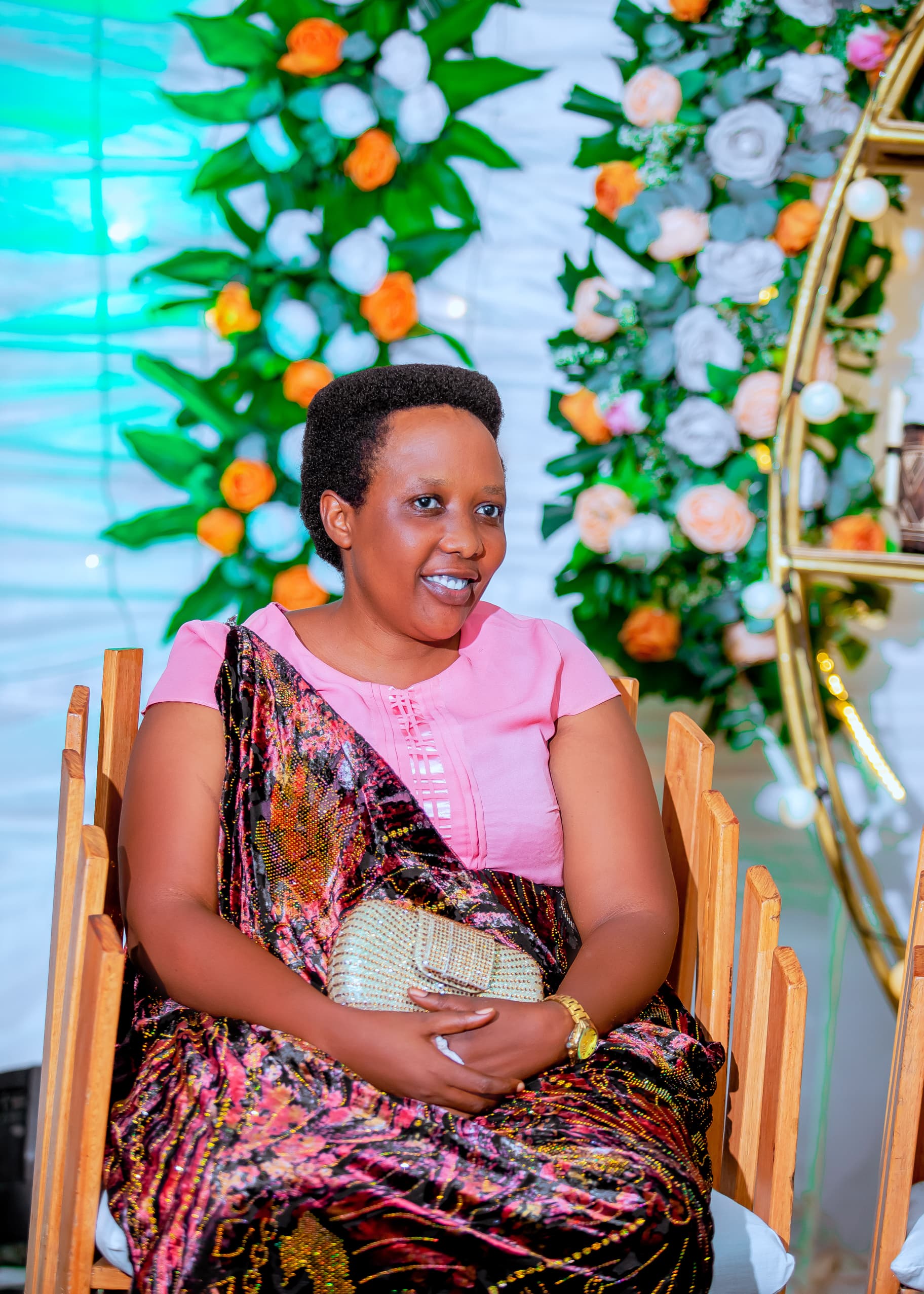 Woman in pink and a patterned wrap seated on a wooden chair beside floral decorations