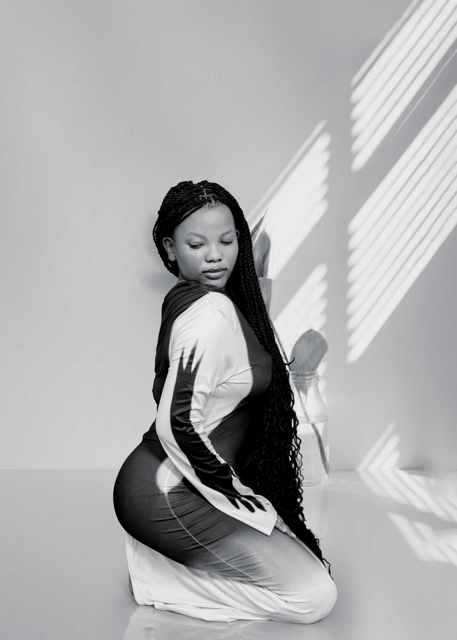Black-and-white portrait of a seated person with long braids looking over their shoulder, with diagonal light and shadows