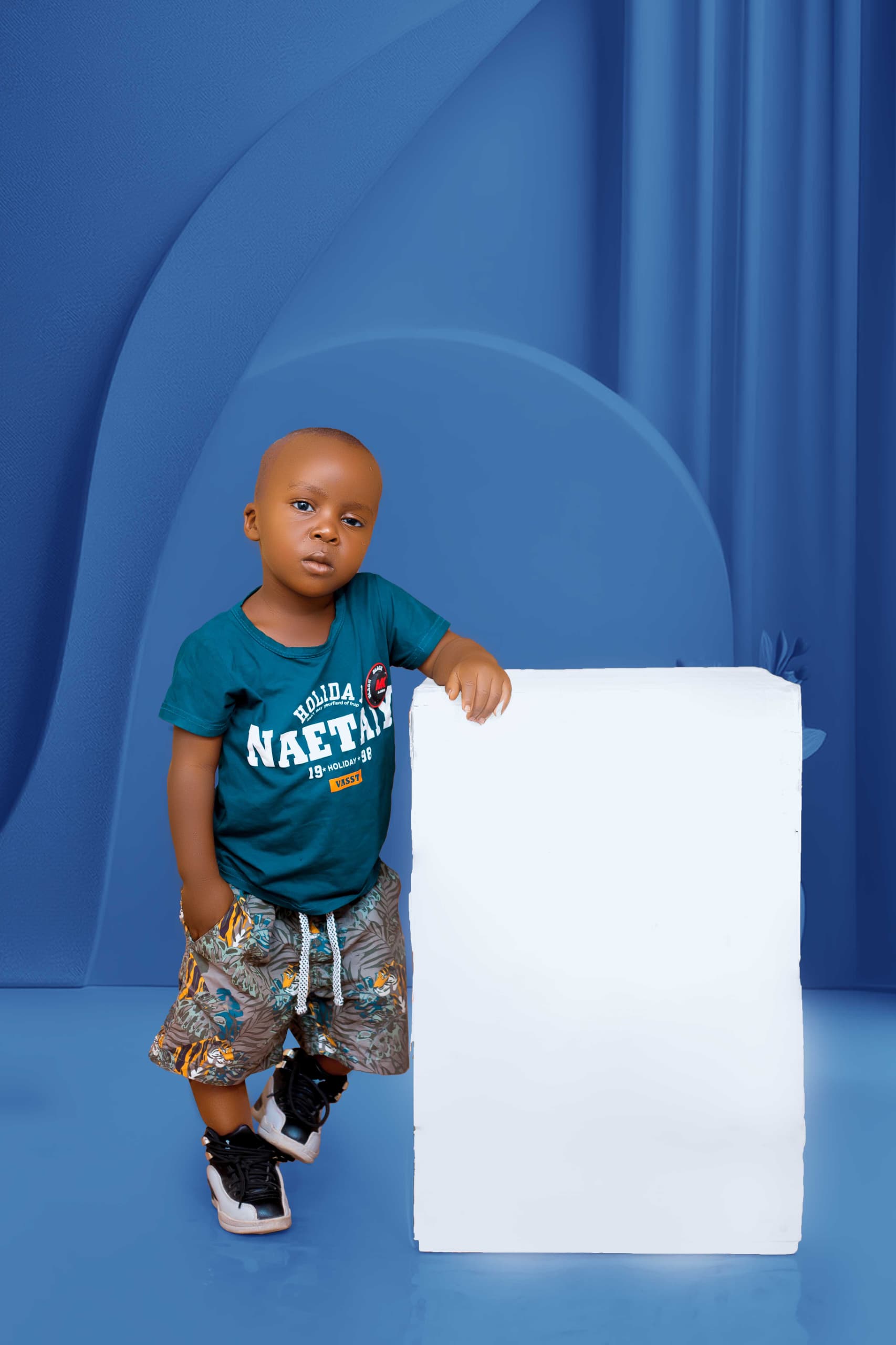 Young child in a teal T-shirt and patterned shorts resting a hand on a white plinth against a blue studio backdrop